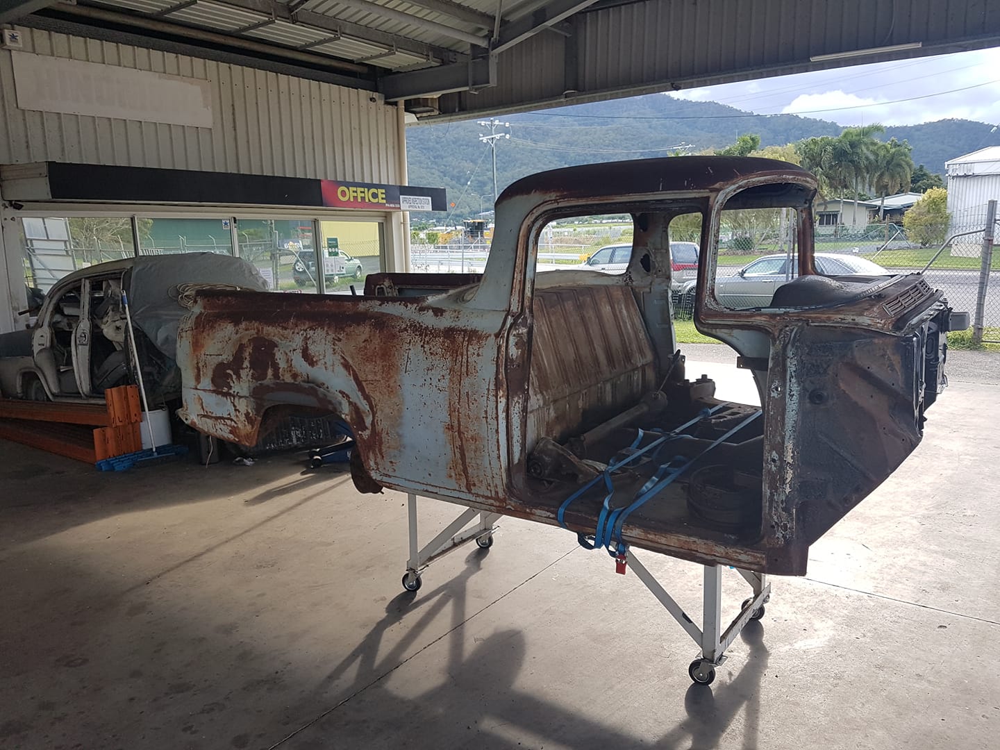 A stripped classic ute cab on a rotisserie in the workshop, with the hills behind
